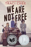 The cover of We Are Not Free. The images of three teens are spraypainted onto side paneling for a house, behind a stack of luggage. The teen on the left has pale skin, blonde hair, and wears a pink dress. The teen in the middle has pale skin, dark hair, and wears a blue overshirt with baggy black pants. The teen on the right has dark hair that covers his eyes, pale skin, and wears a beat up denim jacker over jeans. 
